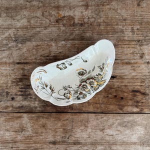 May include: A cream-colored, kidney-shaped dish with floral patterns in shades of brown, gold, and green. The dish has a scalloped edge and is set against a rustic wooden background. A decorative serving piece.