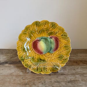 May include: A decorative ceramic plate with a leaf-like border in shades of yellow and green. The plate features two stylized apples in red, yellow, and green. The plate is displayed on a wooden surface.