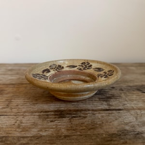 May include: A handmade ceramic candle holder with a floral design. The candle holder is beige with brown floral patterns around the rim. The base is a lighter shade of beige. The candle holder is sitting on a wooden surface.