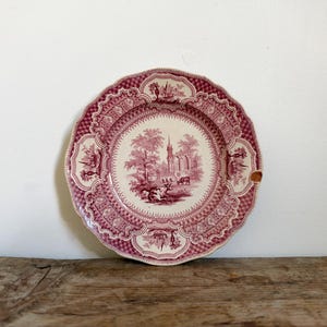 May include: A round ceramic plate featuring a deep red transferware pattern on a cream background. The center depicts a landscape with a church, trees, and cattle. The scalloped rim has a geometric lattice border with small vignettes.