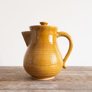 May include: A yellow ceramic pitcher with a striped design. The pitcher has a handle and a lid.