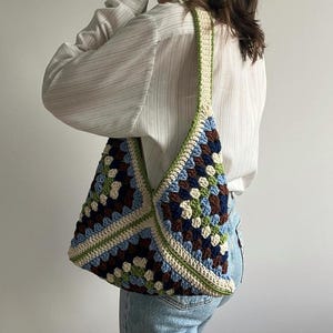 May include: A crochet shoulder bag featuring a granny square pattern in shades of navy blue, light blue, sage green, brown, and cream. The bag has a wide, integrated strap and a triangular silhouette constructed from multiple small yarn clusters.