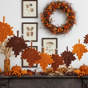 May include: A decorative garland featuring crocheted maple leaves in shades of orange, golden yellow, and dark brown, strung together on a white cord. The leaves are textured with visible yarn stitches and have distinct pointed lobes.