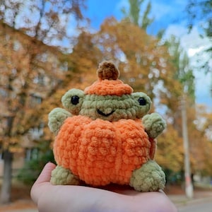 May include: A small, handmade crochet plush toy shaped like a frog wearing a bright orange pumpkin costume. The frog has light green yarn skin, black plastic eyes, and a stitched smile. A small brown stem sits atop the orange pumpkin hat.
