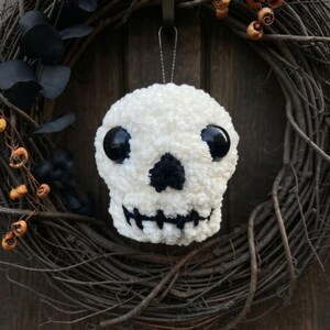 May include: A white crocheted skull ornament with large, round, glossy black plastic eyes, a small black triangular nose, and a stitched black mouth. The skull is suspended by a thin silver string, featuring a textured, soft yarn construction.