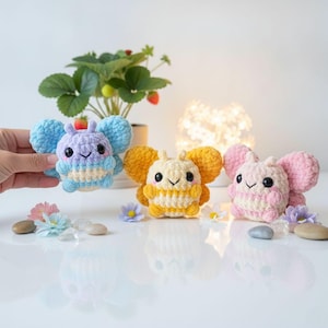 May include: Three small, round, crocheted plush toys shaped like bees with butterfly wings. One is blue and light purple, one is yellow and cream, and one is pink and cream. Each has black eyes, a stitched smile, and pink cheeks.