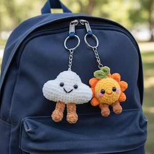 May include: Two crochet keychains attached to a navy blue backpack. One is a white cloud with brown legs and a smiling face. The other is an orange flower with green leaves, brown legs, and a smiling face with pink cheeks. Both have metal rings.