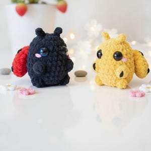 May include: Two small, round, crocheted plush figures with large black eyes and pink stitched cheeks. One figure is black with red wings, and the other is yellow with yellow wings. Both have small antennae and textured yarn bodies.