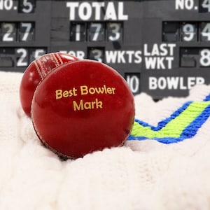 Pode incluir: Duas bolas de críquete de couro vermelho escuro com costuras brancas. Uma bola apresenta o texto gravado em dourado "Best Bowler Mark". Elas repousam sobre um tecido de malha branco com detalhes em azul e verde-limão, diante de um placar.