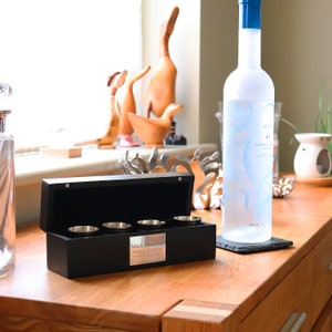 May include: A black wooden box with a silver plaque that reads "Mark Robinson Happy Birthday!" The box is open and contains four silver shot glasses. A bottle of vodka is on a black slate coaster to the right of the box.