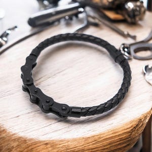Op de afbeelding: Zwart leren armband met een zwart metalen kettingdetail. De armband is gevlochten en heeft een eenvoudige sluiting.