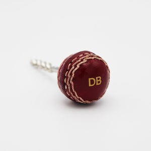 May include: A red leather cricket ball with white stitching and the letters "DB" in gold on the side. The ball is attached to a silver metal key chain.