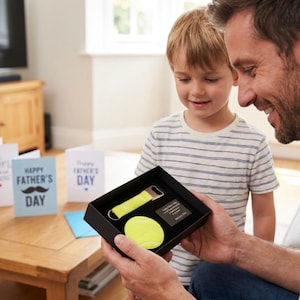 Könnte beinhalten: Eine schwarze Geschenkbox mit einem Untersetzer in Form eines Tennisballs, einem Flaschenöffner mit gelbem Griff und einer kleinen Plakette. Die Box wird von einer Person gehalten, mit einem Kind, das zuschaut. Vatertagskarten sind im Hintergrund zu sehen.