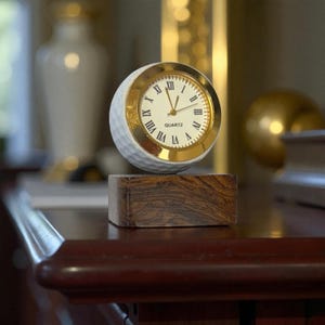 Puede incluir: Un reloj de cuarzo con forma de pelota de golf con un bisel dorado y números romanos. El reloj está sobre un bloque de madera oscura. La esfera del reloj dice "QUARTZ". La pelota de golf tiene una textura con hoyuelos.