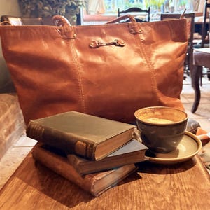 May include: A brown leather tote bag with a metal clasp sits on a wooden table next to a stack of three books and a cup of coffee.