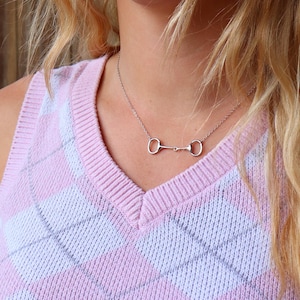 May include: A silver necklace with a horse bit pendant. The necklace is worn by a person wearing a pink and white argyle sweater.