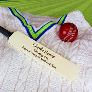 May include: A cricket-themed gift set. It includes a wooden cricket bat with personalized text, a red cricket ball, and a white cable-knit cricket sweater with green and blue trim. The bat has the text "Charlie Harris" and "Happy birthday from Mark and Claire".