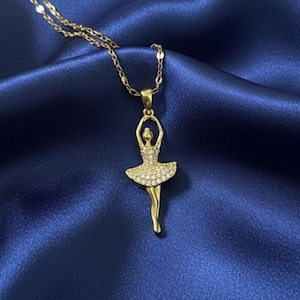 May include: A gold-colored necklace with a pendant shaped like a ballerina. The ballerina's tutu is encrusted with small, sparkling stones. The necklace chain is delicate, and the background is a deep blue fabric.