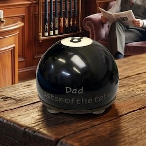 Könnte beinhalten: Eine schwarze Billardkugel mit einem weißen Kreis und der Nummer acht auf der Oberseite. Die Kugel ist auf der Vorderseite mit dem Text "Dad" und "Master of the table" graviert. Es ist ein kugelförmiges Objekt mit glatter, glänzender Oberfläche.