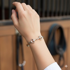 Könnte beinhalten: Silberfarbenes Armband im Reitstil. Das Armband hat ein Gebiss-Design, mit einem zentralen Verbindungssteg und abgerundeten Enden. Das Armband wird am Handgelenk einer Person getragen, mit einem verschwommenen Hintergrund eines Stalls.
