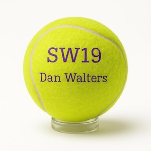 Peut inclure: Une balle de tennis jaune vif avec le texte "SW19" et "Dan Walters" imprimé en violet. La balle repose sur un support transparent et circulaire. La balle a une texture duveteuse et des coutures blanches.