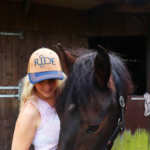 May include: A tan and navy blue trucker hat with the word "RIDE" in navy blue script. The hat is worn by a person with blonde hair, standing next to a dark brown horse. The background is a wooden stable.