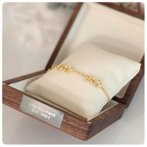 May include: A gold chain bracelet with repeating circular links resting on a white velvet cushion inside a dark wood box. A silver metal plaque on the front of the box is engraved with the text Lily Rochard Est. 2001 in a clean font.