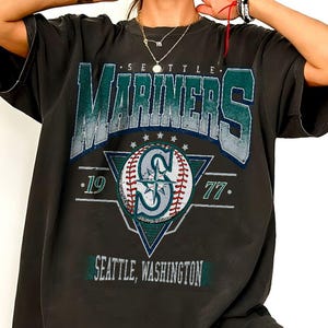 Pode incluir: Uma t-shirt de manga curta cinzento-carvão com uma impressão gráfica que diz Seattle Mariners, com os números 19 e 77 em ambos os lados de um ícone de basebol centrado dentro de um triângulo. O texto Seattle, Washington aparece na parte inferior.