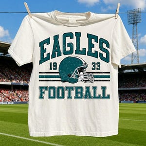 Peut inclure: Un t-shirt à manches courtes de couleur crème avec un motif bleu sarcelle foncé vieilli portant le texte EAGLES 19 33 FOOTBALL et une illustration de casque de football américain au centre, sur fond de stade.