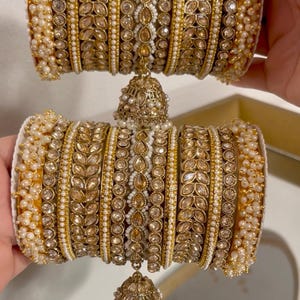 May include: Gold-toned bangles with intricate detailing. The bracelets feature rows of small pearls, clear and amber-colored stones, and ornate metalwork. A dangling bell-shaped charm with pearls hangs from the center. The jewelry is displayed against a neutral background.