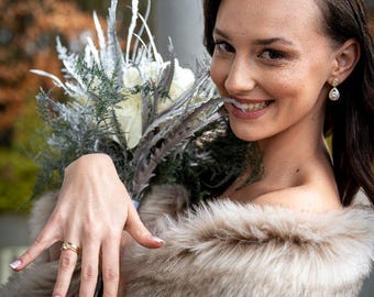 May include: A woman wearing a beige faux fur shawl holds a bouquet of white and silver flowers. She is smiling and wearing a gold ring on her finger.