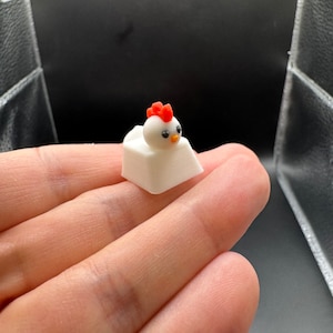 May include: A white chicken keycap with a red comb and black eyes. The keycap is sitting on a hand.