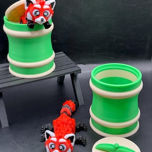 Può includere: Un giocattolo di panda rosso stampato in 3D con un contenitore abbinato verde e crema. Il panda ha un corpo rosso, viso bianco e zampe nere. Il contenitore è cilindrico con coperchio e manico verde.
