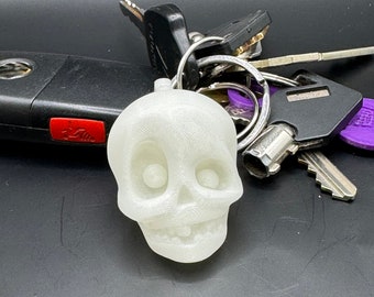 May include: A white glow-in-the-dark skull keychain with a toothy grin. The keychain is attached to a silver ring with a set of keys.