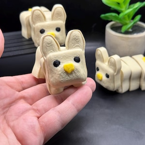 以下が含まれることがあります： 黄色い鼻と黒い目をした、漫画のウサギの3Dプリントされたフィギュア3つ。ウサギのフィギュアはベージュで、表面にテクスチャがあります。フィギュアは黒い表面の上にあります。