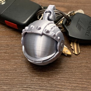 Könnte beinhalten: Ein silberner Astronautenhelm-Schlüsselanhänger mit detailliertem Design. Der Schlüsselanhänger ist an einem Schlüsselsatz befestigt.