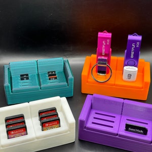 May include: Four 3D printed couch-shaped organizers in teal, orange, white, and purple. The teal organizer has two compartments. The orange organizer has three compartments and holds two USB drives. The white organizer has two compartments and holds Nintendo Switch game cartridges. The purple organizer has two compartments and holds a SD card.