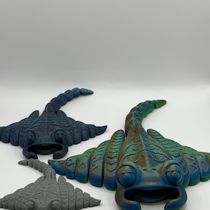 Op de afbeelding: Drie 3D-geprinte roggensculpturen in blauw- en grijstinten. De sculpturen hebben gedetailleerde kenmerken, waaronder ogen, monden en gesegmenteerde staarten. De grootste rog heeft een verloop van blauw en groen.