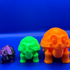 May include: Three 3D printed turtle figurines in different sizes. The largest turtle is orange, the middle turtle is green, and the smallest turtle is purple.