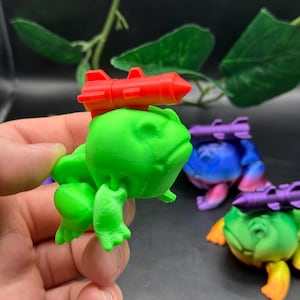 May include: Three green frog figurines with missiles on their heads. The frogs are different shades of green and have a cartoonish appearance. The missiles are red, purple, and blue.