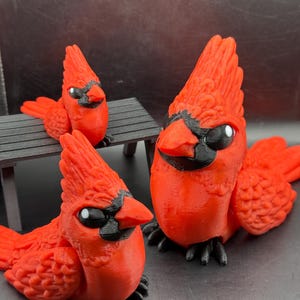 May include: Three red cardinal figurines with black accents. The birds have detailed feather textures, black beaks, and eyes. One cardinal is perched on a small black bench. The figurines are displayed on a dark surface.