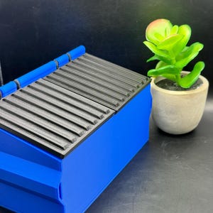 May include: A blue plastic toy dumpster with a black lid. The lid is open and has a ribbed texture. The dumpster is sitting on a black surface.