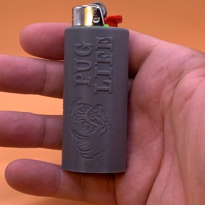 May include: A gray lighter with a custom sleeve featuring the words "Pug Life" and a pug dog illustration. The lighter has a silver metal top, a red ignition button, and a green wheel.