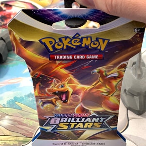 May include: A Pokemon Trading Card Game booster pack featuring a fiery orange and yellow Charizard on the front. The pack is labelled "Sword & Shield - Brilliant Stars" and includes 10 additional game cards.
