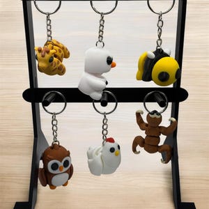 以下が含まれることがあります： 6つのキーホルダーを保持する黒い金属製のディスプレイスタンド。キーホルダーは、キリン、雪だるま、蜂、フクロウ、鶏、ナマケモノなど、動物のデザインが特徴です。各キーホルダーは、銀色の金属リングに取り付けられています。