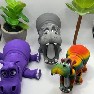 May include: Three 3D printed hippopotamus figurines. One is purple, one is gray, and one is multi-colored with green, yellow, orange, and red.