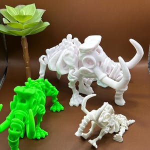 Puede incluir: Tres esqueletos de dinosaurios impresos en 3D, uno verde y dos blancos. Los esqueletos blancos son más grandes que el esqueleto verde.