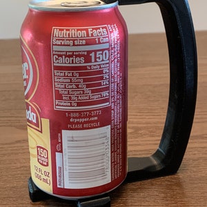 May include: A black 3D printed can holder with a handle. The holder is designed to fit a standard 12-ounce can. The can holder is shown holding a red can of Dr Pepper. The can has the nutrition facts label visible, which states that the can contains 150 calories, 0 grams of fat, 55 milligrams of sodium, 40 grams of carbohydrates, and 39 grams of sugar. The can also has a barcode and the Dr Pepper logo.