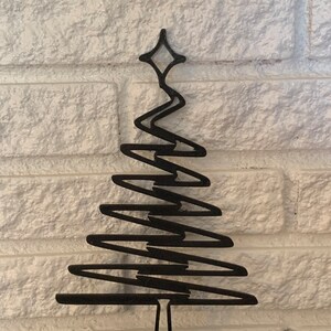 May include: Black metal Christmas tree ornament with a star on top and a zig-zag design. The ornament is on a white brick background.