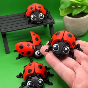 May include: A collection of red and black ladybug figurines with large, cartoonish eyes. One ladybug is held in a hand, while others are arranged on a small black bench and a green surface. The ladybugs have a 3D printed appearance.